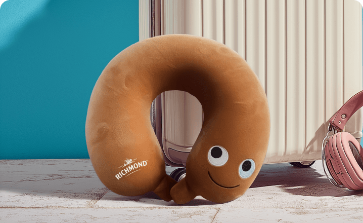 A Richmond-branded sausage-shaped travel neck pillow resting against a suitcase, one of the 250 available prizes.