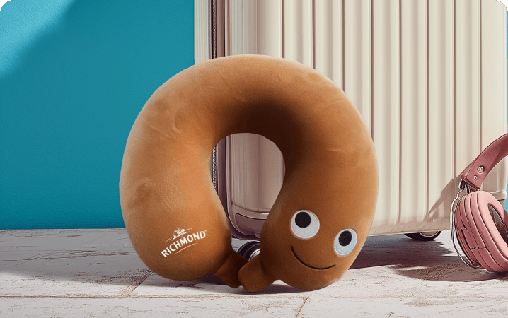 A Richmond-branded sausage-shaped travel neck pillow resting against a suitcase, one of the 250 available prizes.